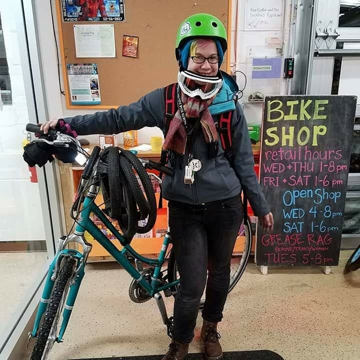 Teal Raleigh commuter road bike with front studded tire, old road tires around handlebar; femme transmasculine winter cyclist with brown boots, jeans, grey jacket, mulitcolor scarf, white goggles around neck, teal handband under lime green helmet; background is inside Cycles for Change with open shop sandwich board hours behind cyclist to the right.